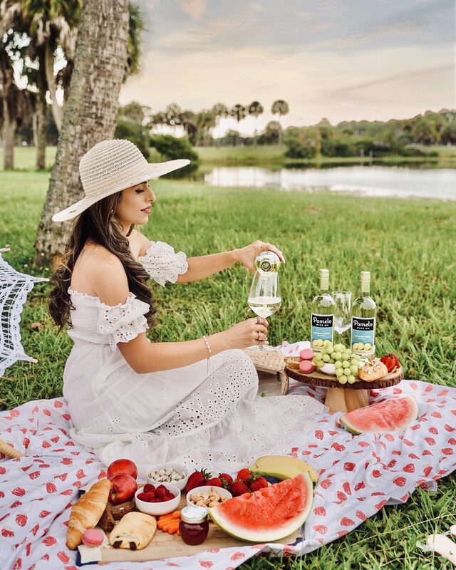 Roopsi Bansal in a white ruffled off-shoulder eyelet dress and straw hat, pouring a glass of Pomelo Wines Sauvignon Blanc at a picnic spread of fruit and pastries beside a palm-lined lake