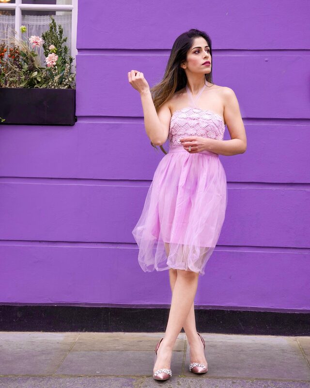 Roopsi Bansal in a pink lace-bodice tulle dress, standing against a bold violet-painted shopfront with a window box of pink flowers