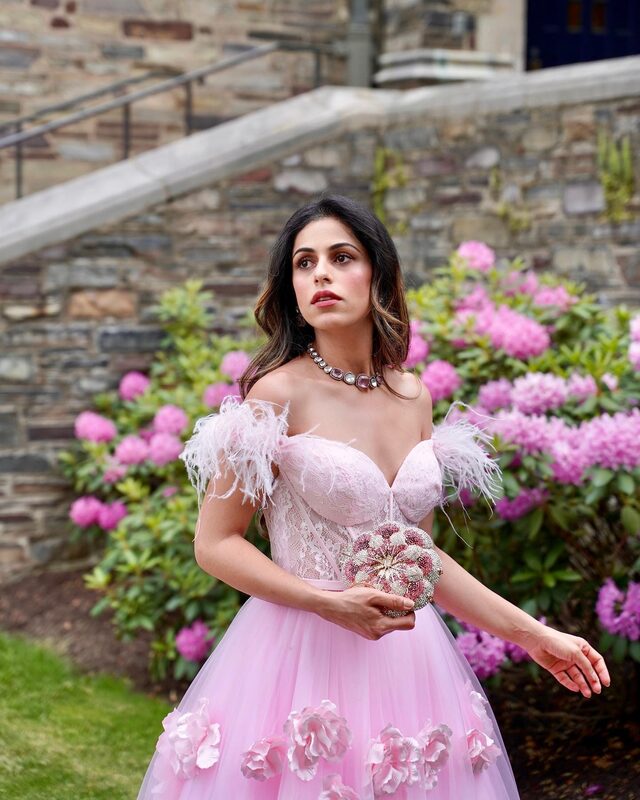 Roopsi Bansal in a pink feather-trim ballgown with 3D floral appliques standing in front of blooming pink azalea bushes