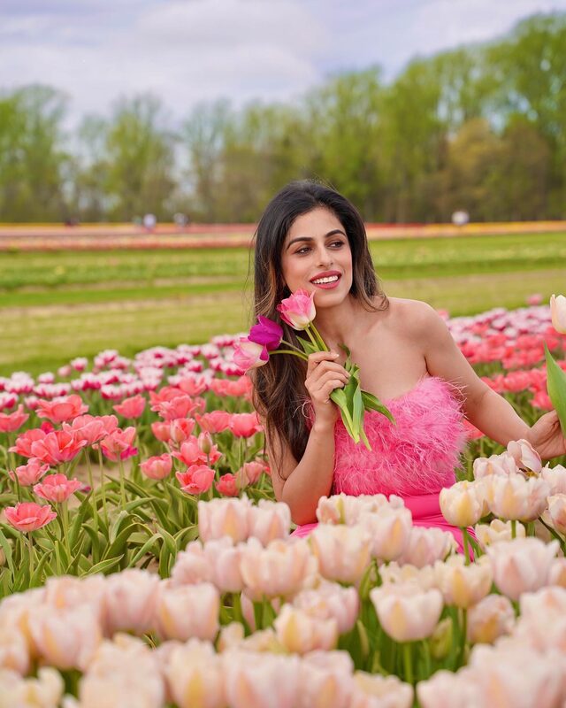 Roopsi Bansal in a pink feather-trim dress sitting among rows of pink and cream tulips holding a small bouquet