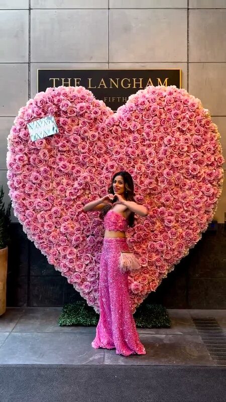 Roopsi Bansal in a pink sequin crop top and skirt making a heart shape with her hands in front of a giant heart made of pink roses at The Langham