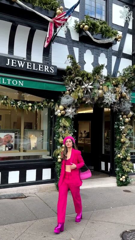 Roopsi Bansal in a hot pink lace pantsuit and beret in front of a jeweler's storefront decorated with garland and ornaments