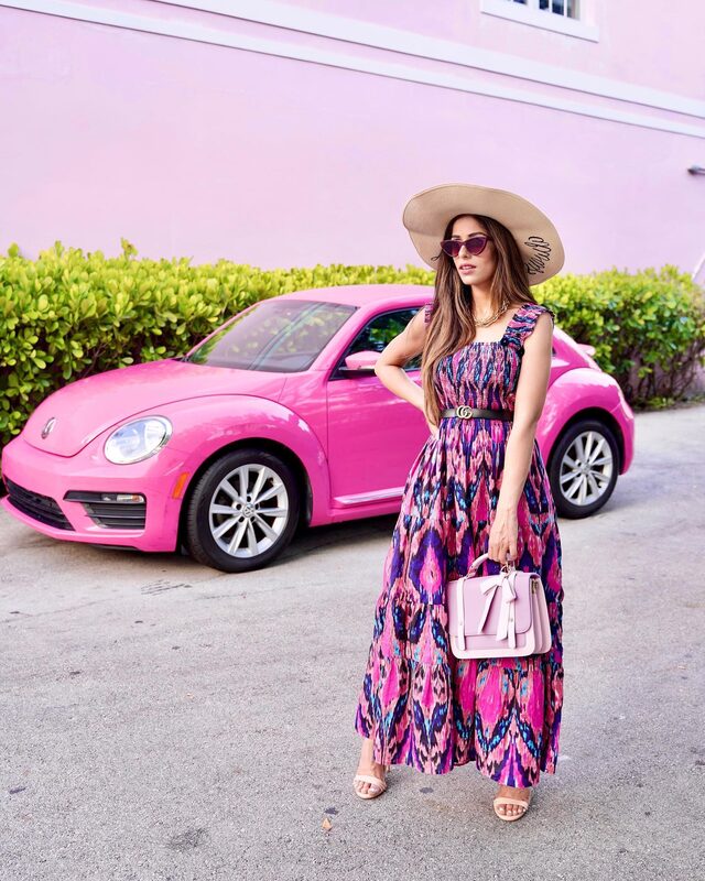 Roopsi Bansal in a pink and purple ruffled maxi dress and a wide straw hat, standing in front of a hot pink Volkswagen Beetle parked outside Big Pink diner in Miami