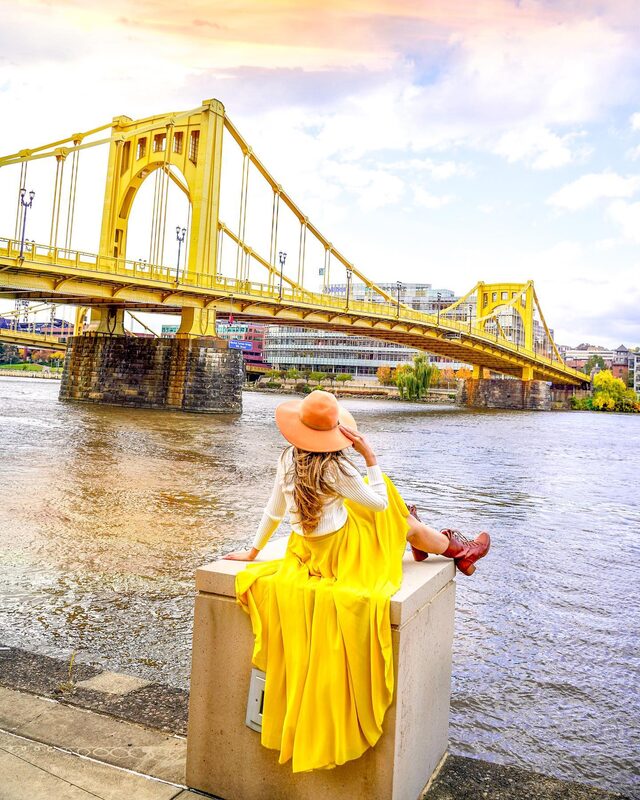 Roopsi Bansal from behind in a flowing yellow maxi skirt and orange floppy hat, seated on a stone ledge facing the yellow Roberto Clemente Bridge over the Allegheny River in Pittsburgh