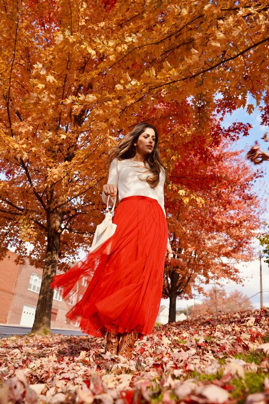 Roopsi Bansal in a cream knit top and flowing red tulle skirt, standing among red and gold fall leaves on Mt. Washington in Pittsburgh