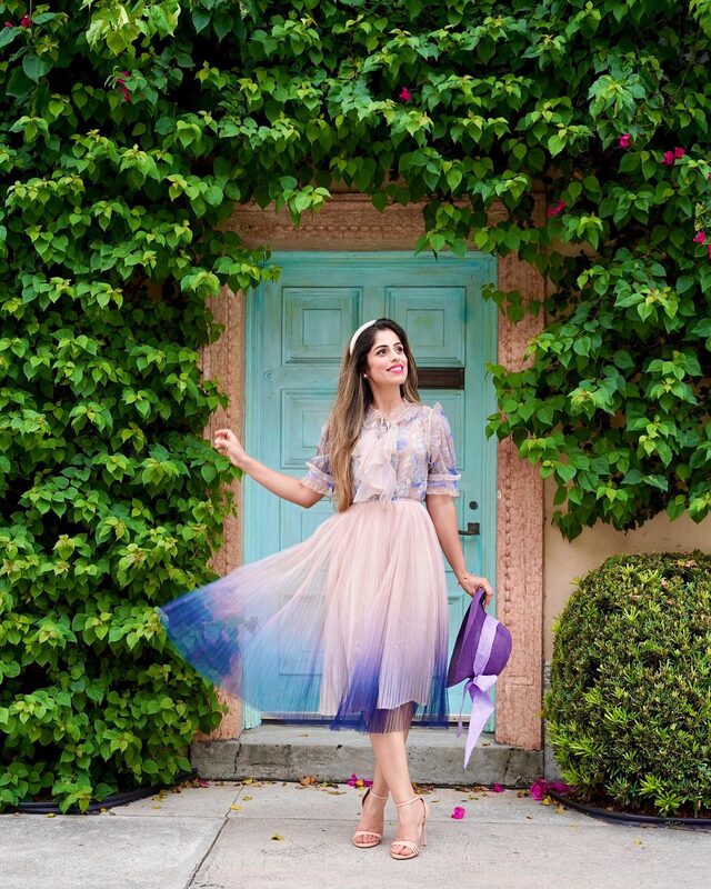 Roopsi Bansal in a sheer floral blouse and an ombre pink-to-blue pleated tulle skirt, standing in front of a vine-covered turquoise door in West Palm Beach