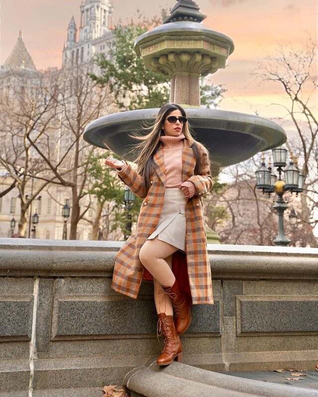 Roopsi Bansal in a plaid coat and cream skirt leaning against an ornate fountain near City Hall Park at golden hour