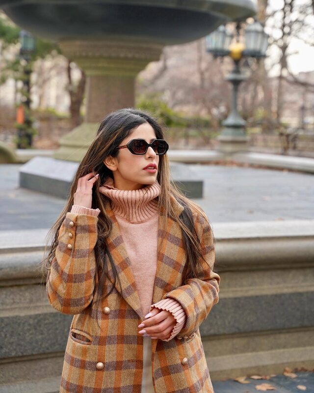 Roopsi Bansal in dark sunglasses and a plaid coat in front of an ornate fountain in City Hall Park