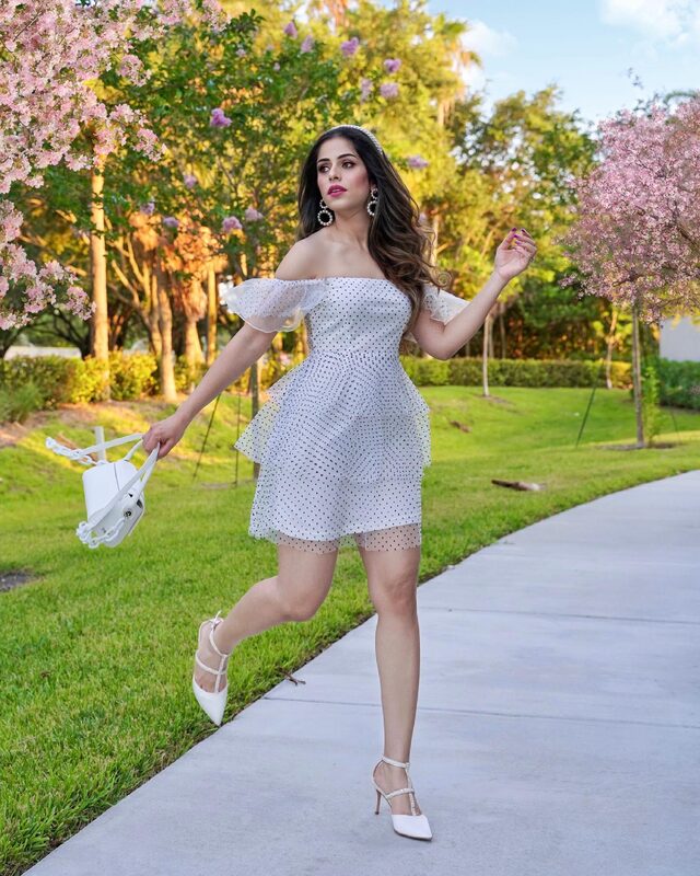 Roopsi Bansal skipping in a white polka-dot ruffle dress and pearl headband, holding a white handbag, under trees in pink blossom