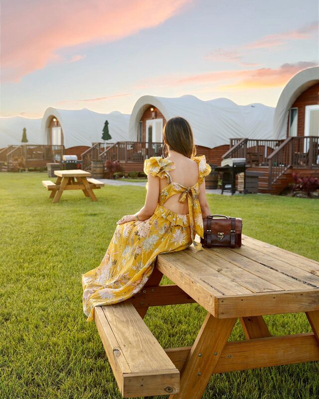 Roopsi Bansal from behind in a yellow floral open-back maxi dress, seated on a picnic table facing a row of wagon-shaped glamping tents at Westgate River Ranch at sunset