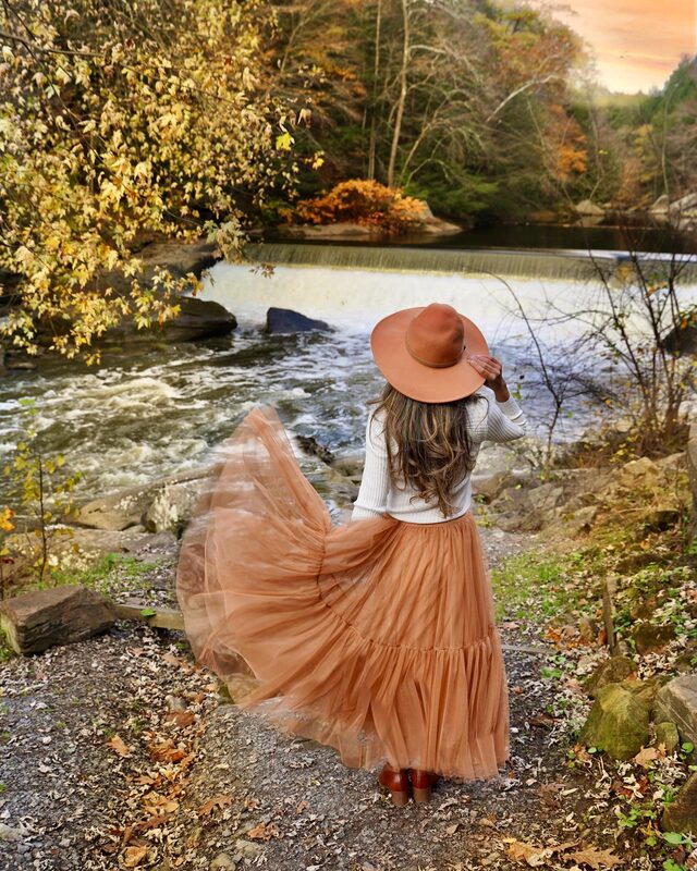Roopsi Bansal from behind in a camel mesh tulle skirt and floppy hat, facing a waterfall surrounded by fall foliage at McConnells Mill State Park