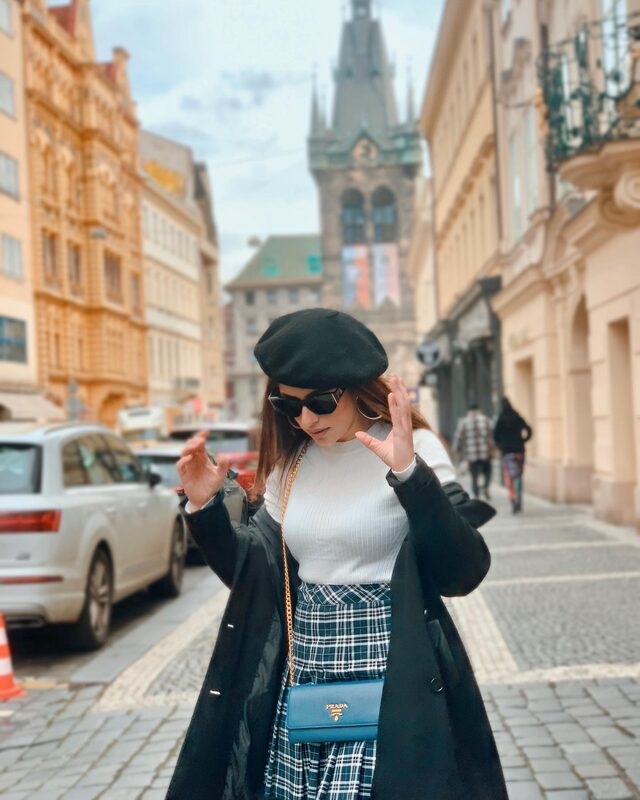 Standing on a cobblestone Prague street with a Gothic tower behind, wearing a black beret, sunglasses, a white top, plaid pleated skirt, and a blue Prada crossbody bag, hands raised playfully