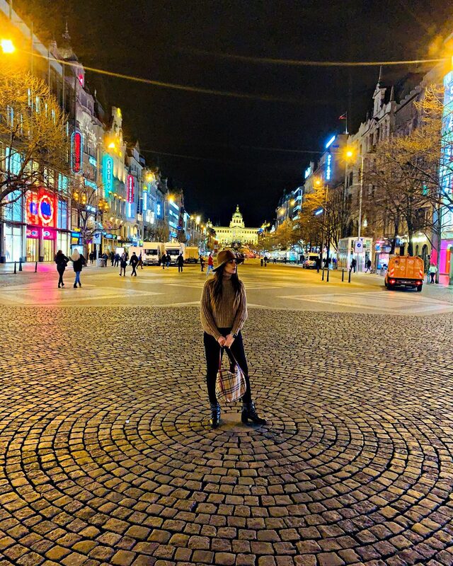 Standing in the middle of a large cobblestone square at night, wearing a brown hat and sweater, holding a Burberry bag, with neon shop signs and a golden-lit building glowing in the distance