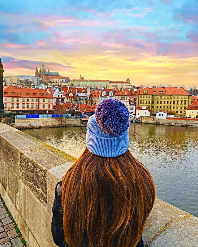 Back-of-head view on Charles Bridge, wearing a purple pom-pom beanie, looking across the river toward Prague Castle lit up in pink and gold sunset light