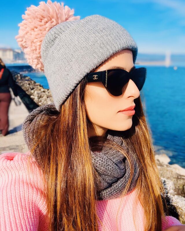 A close-up profile portrait wearing Chanel sunglasses, a grey pom-pom beanie, and a pink sweater, sitting on rocks beside bright blue water