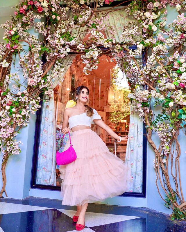 Roopsi Bansal in a white one-shoulder top and a blush layered tulle skirt, holding a hot pink handbag in front of a storefront covered floor to ceiling in pink and white blossoms