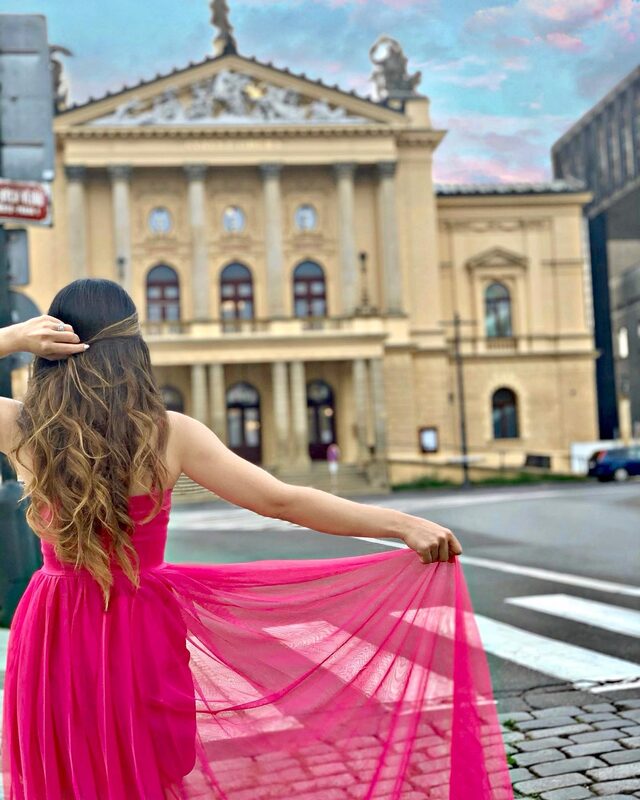 Roopsi Bansal in a flowing hot-pink gown, back to camera, facing a grand columned theatre building in Prague at dusk