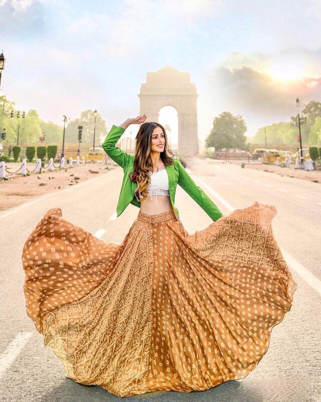 Roopsi Bansal twirling in a gold bandhani skirt, white lace crop top, and green blazer, with India Gate in Delhi glowing in the morning sun behind her