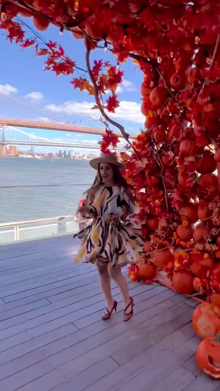 Roopsi Bansal in a black, white, and yellow print dress and straw hat beside the Pumpkin Arch at Pier 17, Brooklyn Bridge in the background