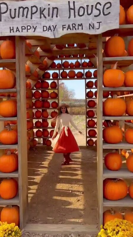 Roopsi Bansal in a white top and red pleated skirt walking through a doorway stacked floor to ceiling with pumpkins at the Pumpkin House, Happy Day Farm NJ