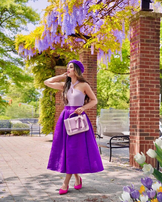 Roopsi Bansal in a purple beret and purple jacquard midi skirt standing under a brick pergola draped in wisteria