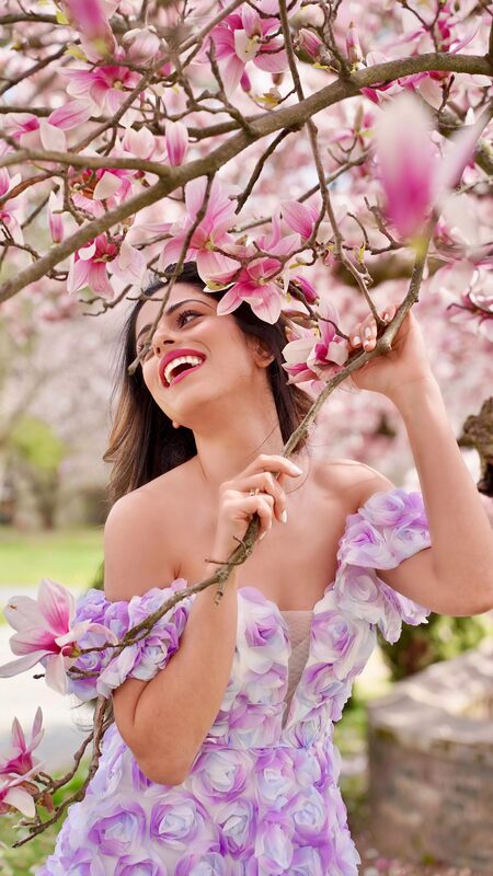 Roopsi Bansal laughing under a magnolia branch in a purple 3D floral off-shoulder dress