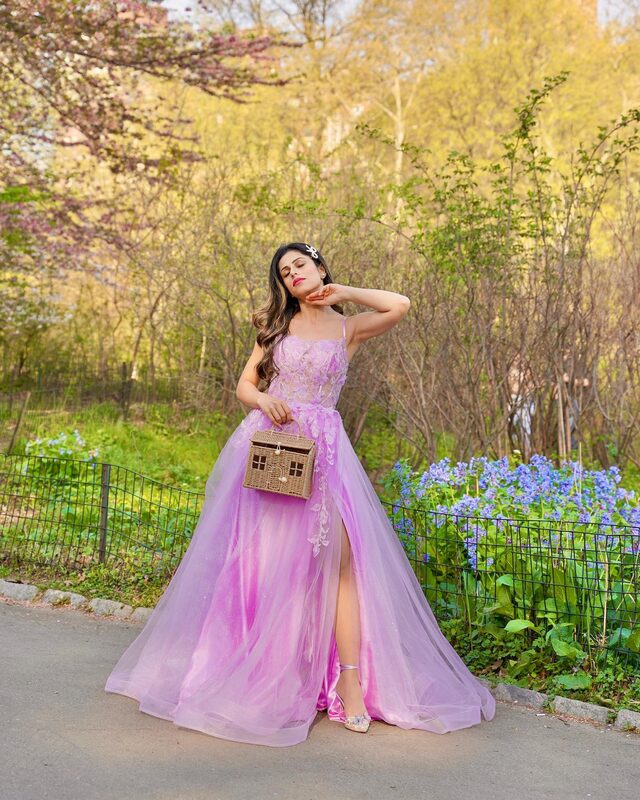 Roopsi Bansal in a lilac tulle gown with a house-shaped straw bag standing on a path in Central Park surrounded by spring greenery