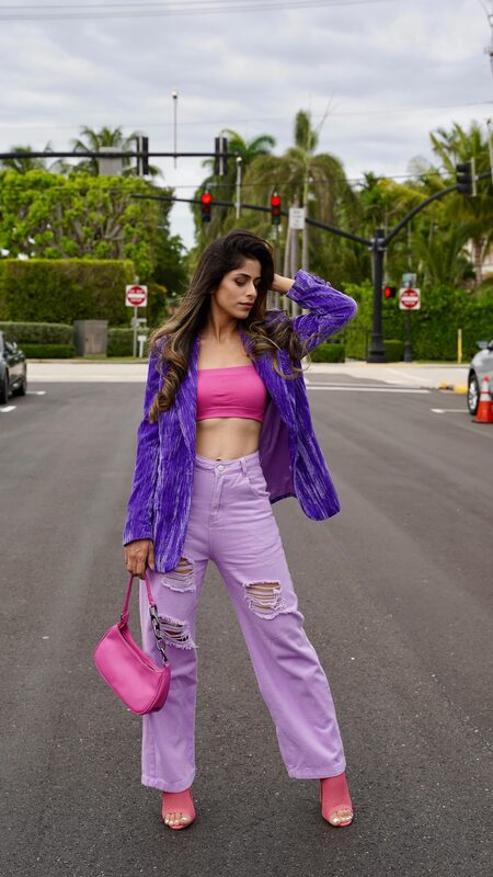 Roopsi Bansal in a purple velvet blazer over a hot pink crop top and lilac ripped straight-leg jeans, holding a pink shoulder bag, standing in the middle of a palm-lined intersection