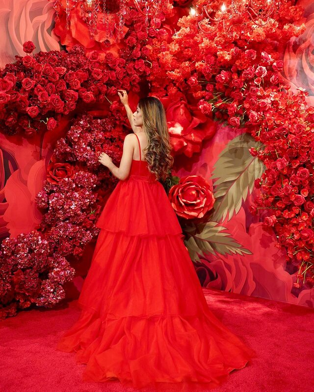 Roopsi Bansal from behind in a tiered red ball gown, reaching toward a wall entirely covered in red roses and hydrangeas, a small red chandelier overhead
