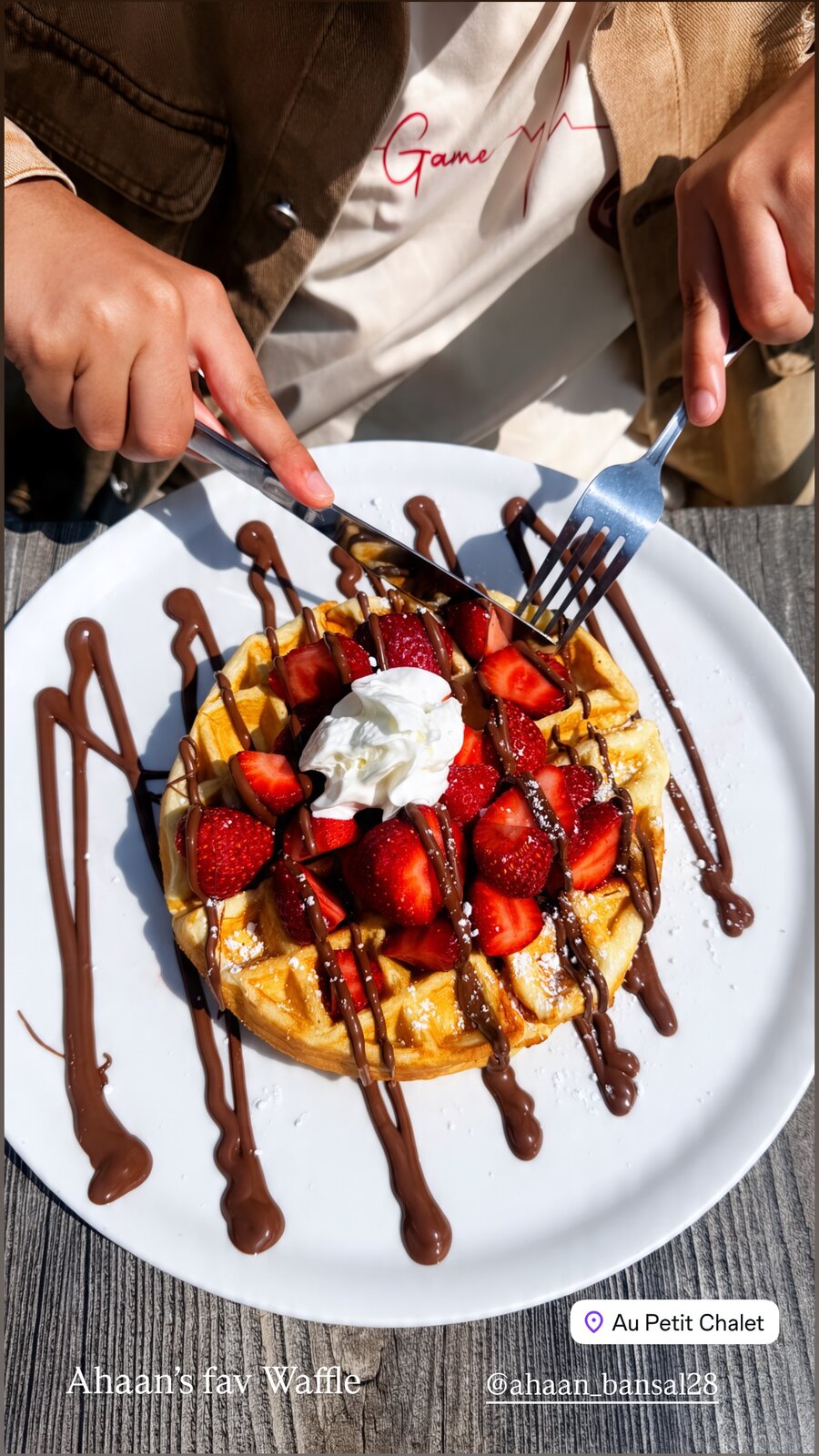 A chocolate-drizzled waffle topped with strawberries and whipped cream being cut into at Au Petit Chalet, captioned Ahaan's fav Waffle