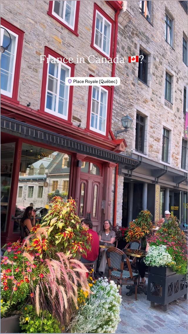 Stone buildings with red-trimmed windows and overflowing flower boxes on a cafe patio at Place Royale in Quebec City, captioned France in Canada