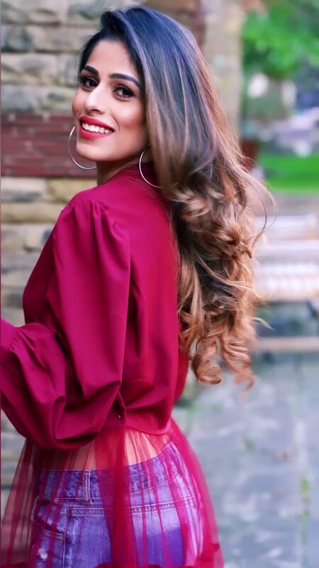 Roopsi Bansal in a berry-red puff-sleeve blouse over a sheer skirt and denim shorts, glancing back over her shoulder against a stone wall