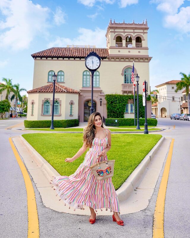 Roopsi Bansal in a rainbow candy-stripe tasselled maxi dress, carrying a straw tote, in front of the Palm Beach Town Hall and its landmark clock tower