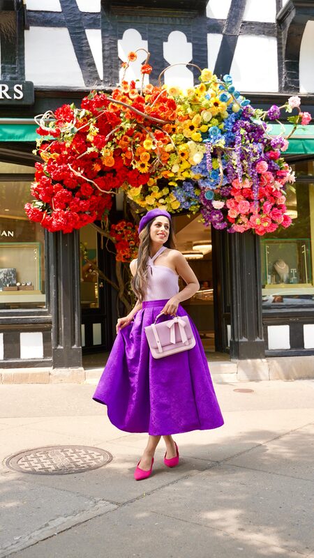 Roopsi Bansal in a purple beret and purple skirt standing beneath a rainbow-colored floral canopy outside a Tudor-style storefront