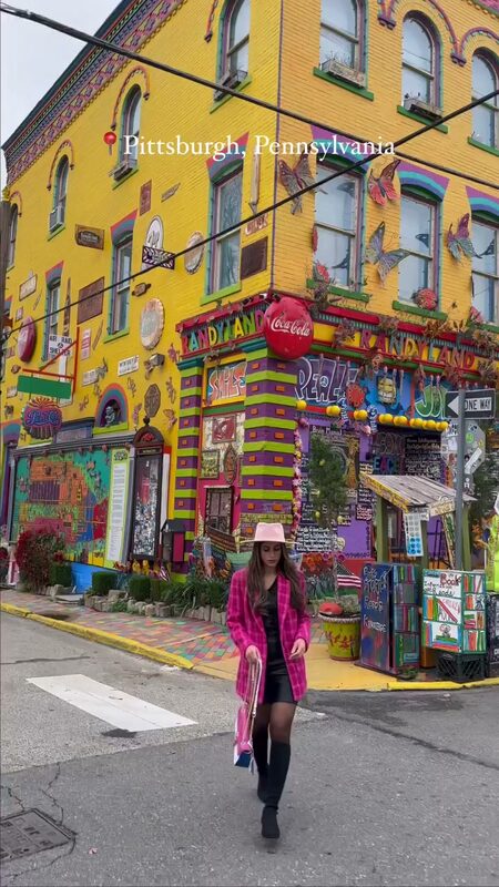 Roopsi Bansal in a pink plaid blazer and pink hat walking in front of the colorful painted murals of Randyland in Pittsburgh