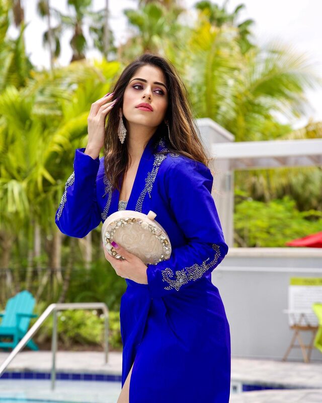Roopsi Bansal in a sapphire blue silk jacket dress with crystal embroidery, holding a jewelled clutch beside a poolside garden