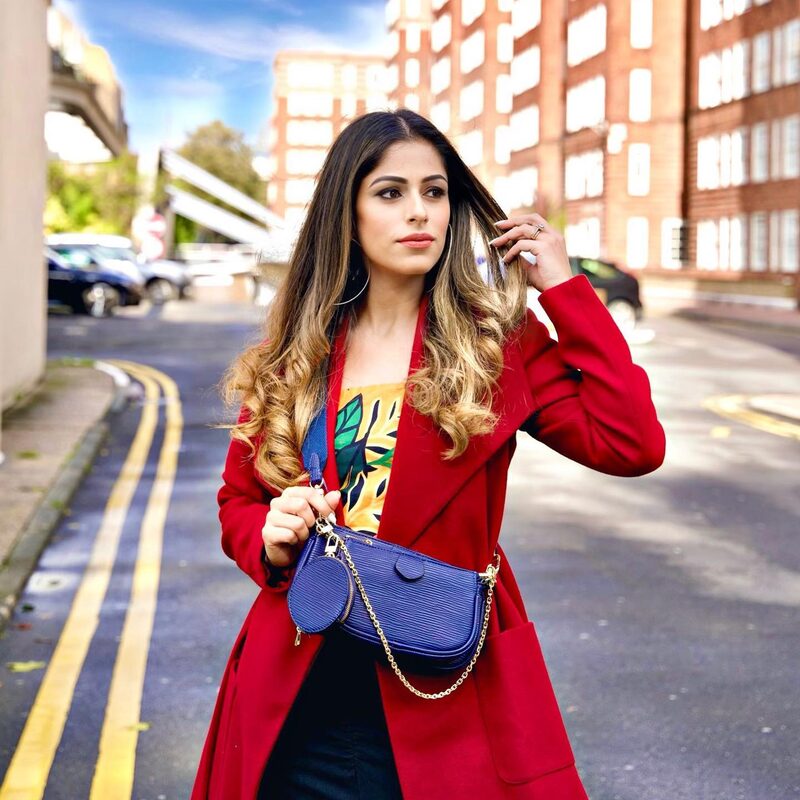 Roopsi Bansal in a red coat over a tropical-print top, carrying a small blue crossbody bag, standing on a London street lined with red-brick buildings
