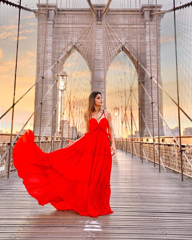 Roopsi Bansal in a flowing red gown on the Brooklyn Bridge walkway at sunset, the dress caught mid-motion in the wind