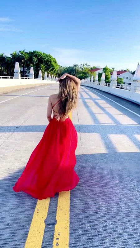 Roopsi Bansal from behind in a flowing red open-back gown, walking across a white footbridge lined with palm trees in Miami