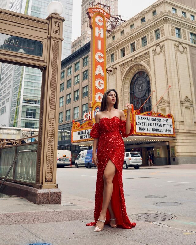 Roopsi Bansal in a sequined red off-shoulder gown with a thigh-high slit, standing on the sidewalk beneath the lit Chicago Theatre marquee