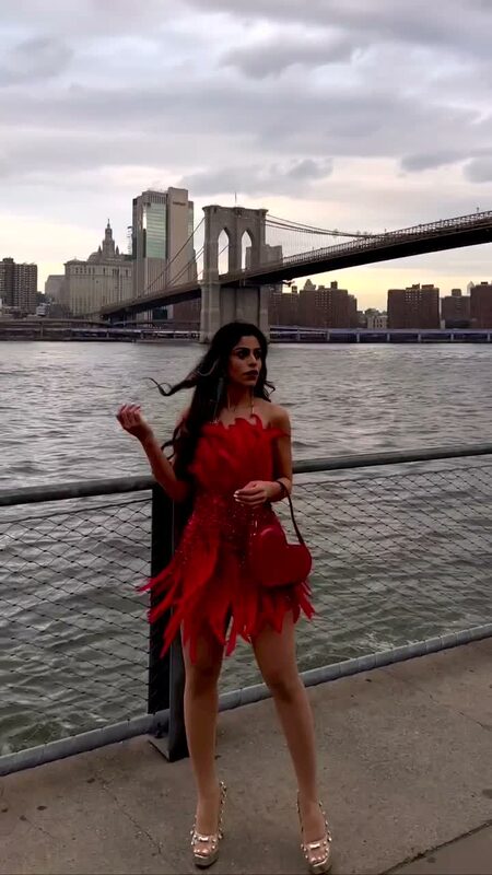 Roopsi Bansal in a red feather mini dress standing on the Brooklyn waterfront at dusk with the Brooklyn Bridge behind her