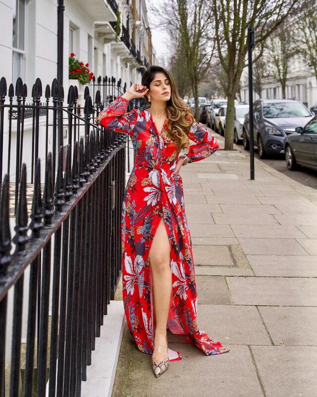 Roopsi Bansal leaning against a black iron railing on a white terraced London street, wearing a red floral maxi dress with a thigh-high slit