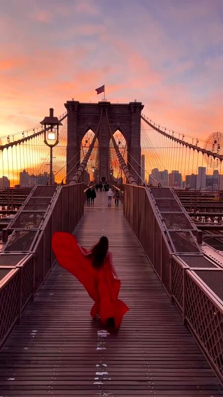 Roopsi Bansal in a flowing red gown walking across the Brooklyn Bridge as the sunrise turns the sky pink and orange