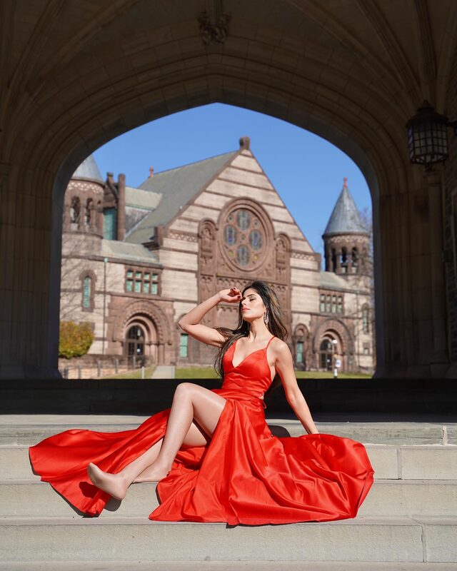 Roopsi Bansal in a red satin gown seated on stone steps under a Gothic stone archway at Princeton, New Jersey