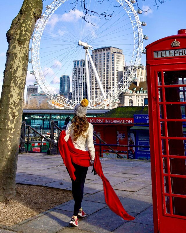Roopsi Bansal from behind in a cream sweater and a red scarf blowing in the wind, walking toward the London Eye and a red telephone box on a sunny day