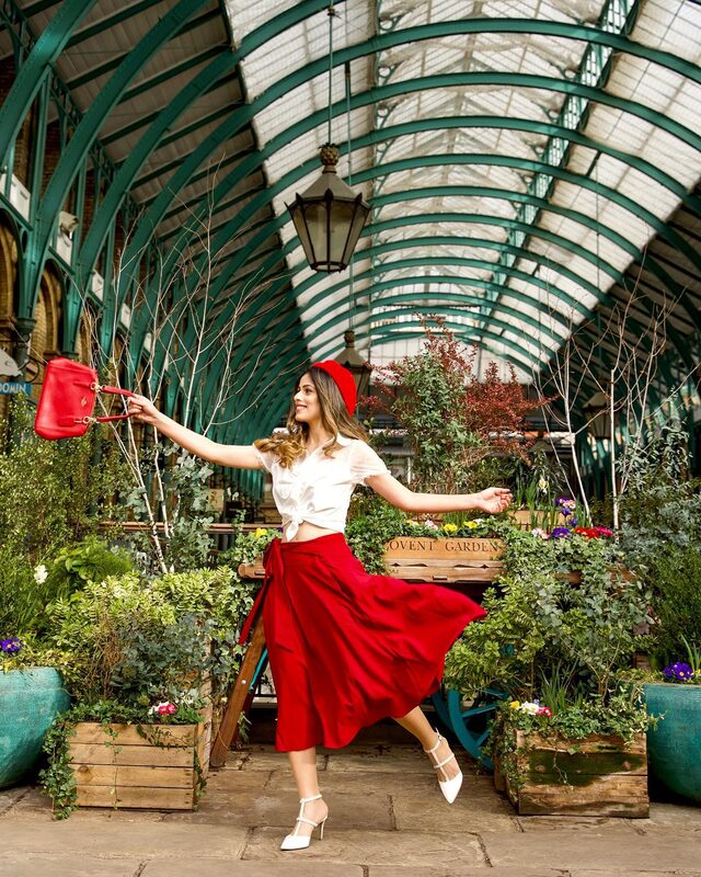 Roopsi Bansal mid-twirl in a red wrap skirt, white blouse, and red beret, swinging a red handbag under the glass-and-iron roof of the Apple Market at Covent Garden