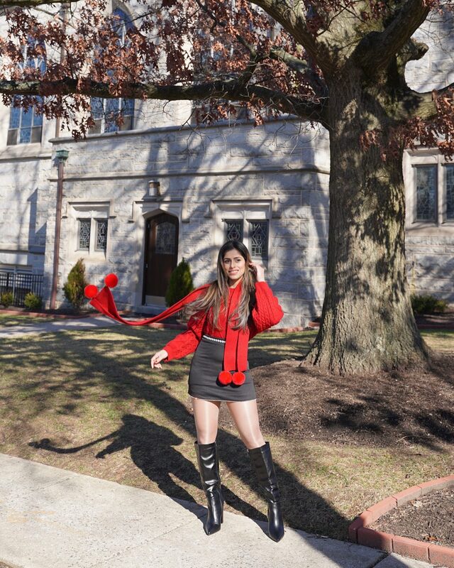Roopsi Bansal in a red cable knit sweater with a pom-pom scarf and black mini skirt jumping in front of a stone building