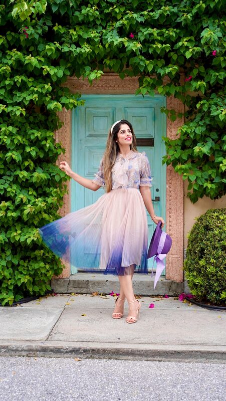 Roopsi Bansal in a lavender ombré tulle skirt and floral top, twirling in front of a vine-covered archway with a turquoise door