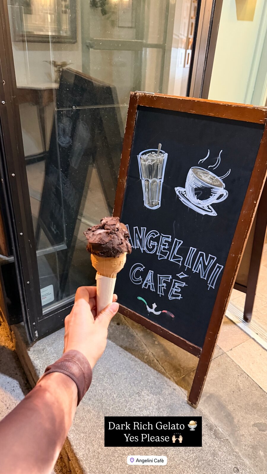 A hand holding a cone of dark chocolate gelato in front of the Angelini Café chalkboard sign