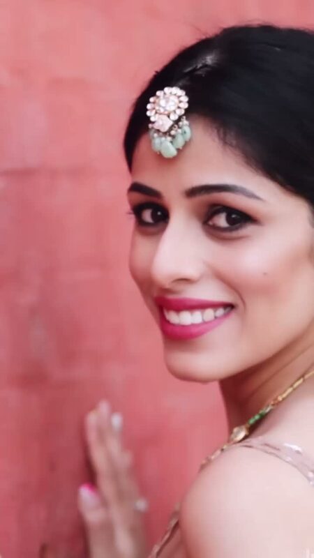 Close-up of Roopsi Bansal wearing a jewelled maang tikka with pink and green gemstones, smiling against a coral pink wall